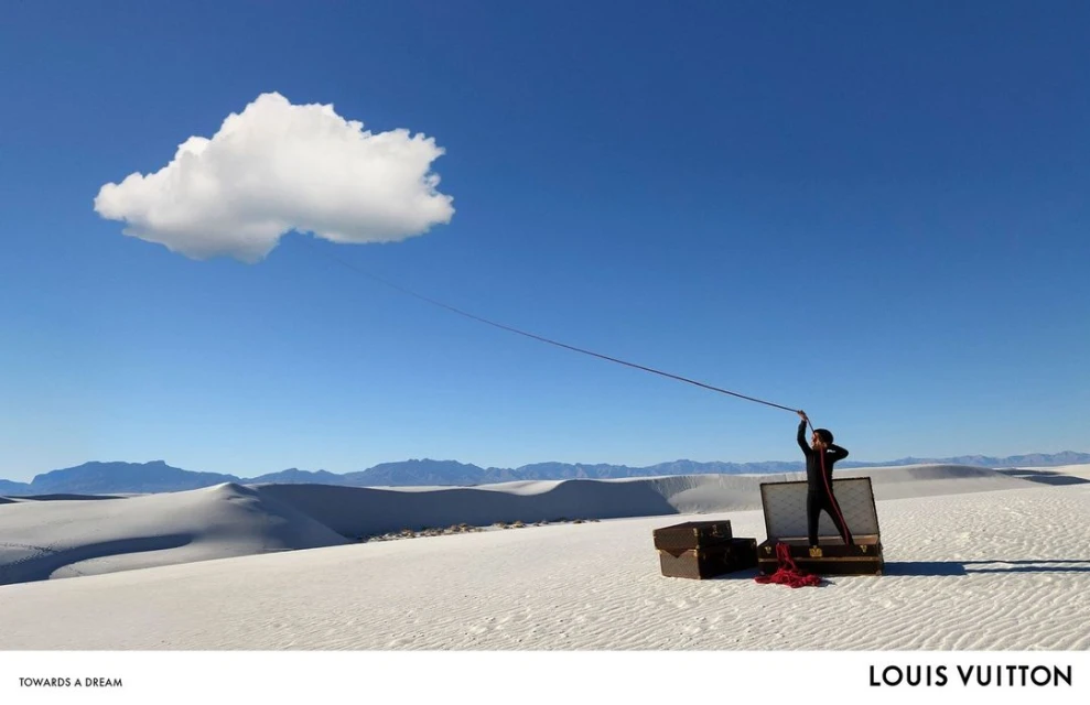Whimsical desert scene: Person stands by open luggage with a red cloth, appearing to hold a string attached to a white cloud in the sky.