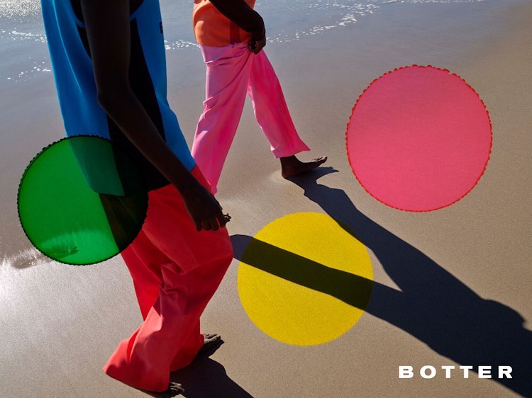 Two people walk barefoot on a sandy beach next to colorful circular shapes in this outdoor fashion shot. Bright sunlight and shadows.