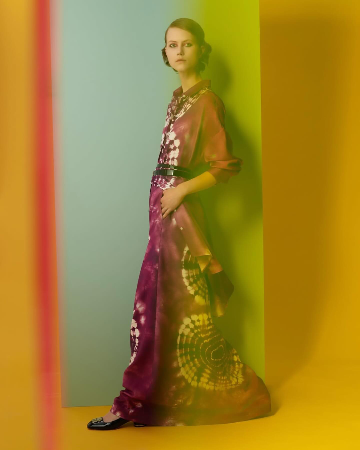 A woman stands in a purple and white dress with a black belt in front of a yellow and blue backdrop, a fashion studio shot.