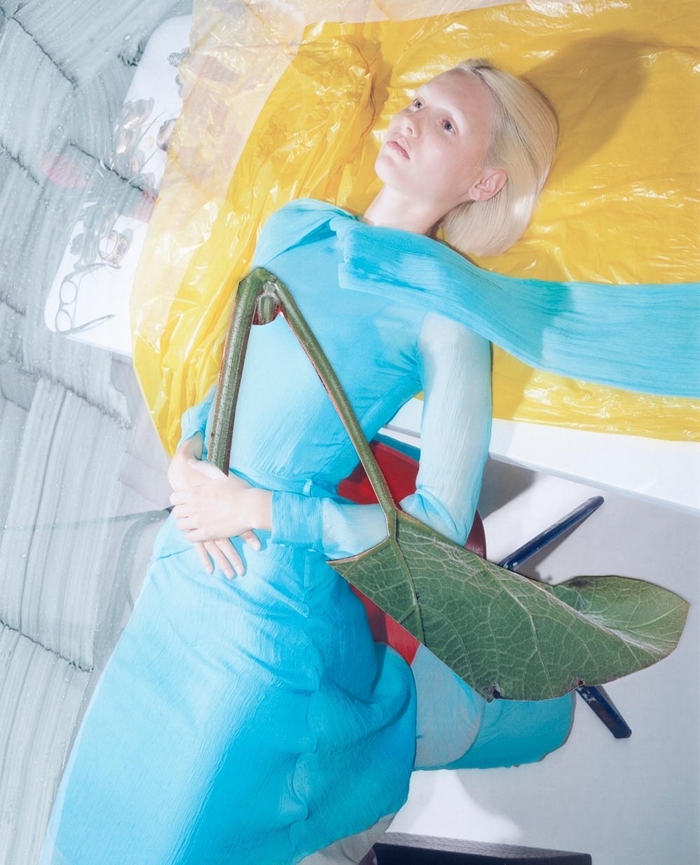 Fashion shot of a blonde model in a blue outfit with a large green leaf accessory, lying against a yellow and grey surface.