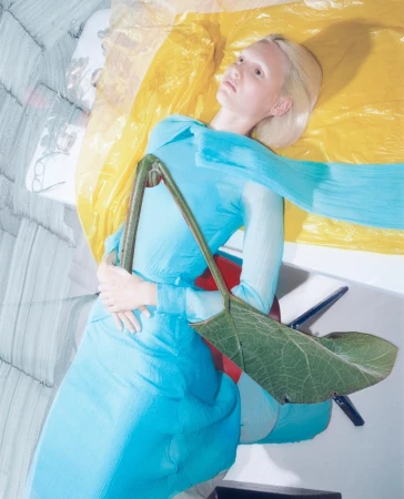 Fashion shot of a blonde model in a blue outfit with a large green leaf accessory, lying against a yellow and grey surface.