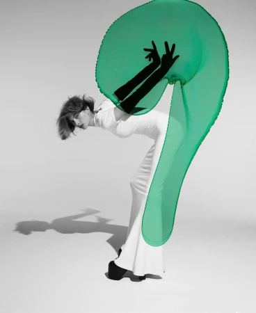 An adult female fashion model poses in a white dress with a large transparent green fabric, captured in a studio with a minimalist aesthetic.