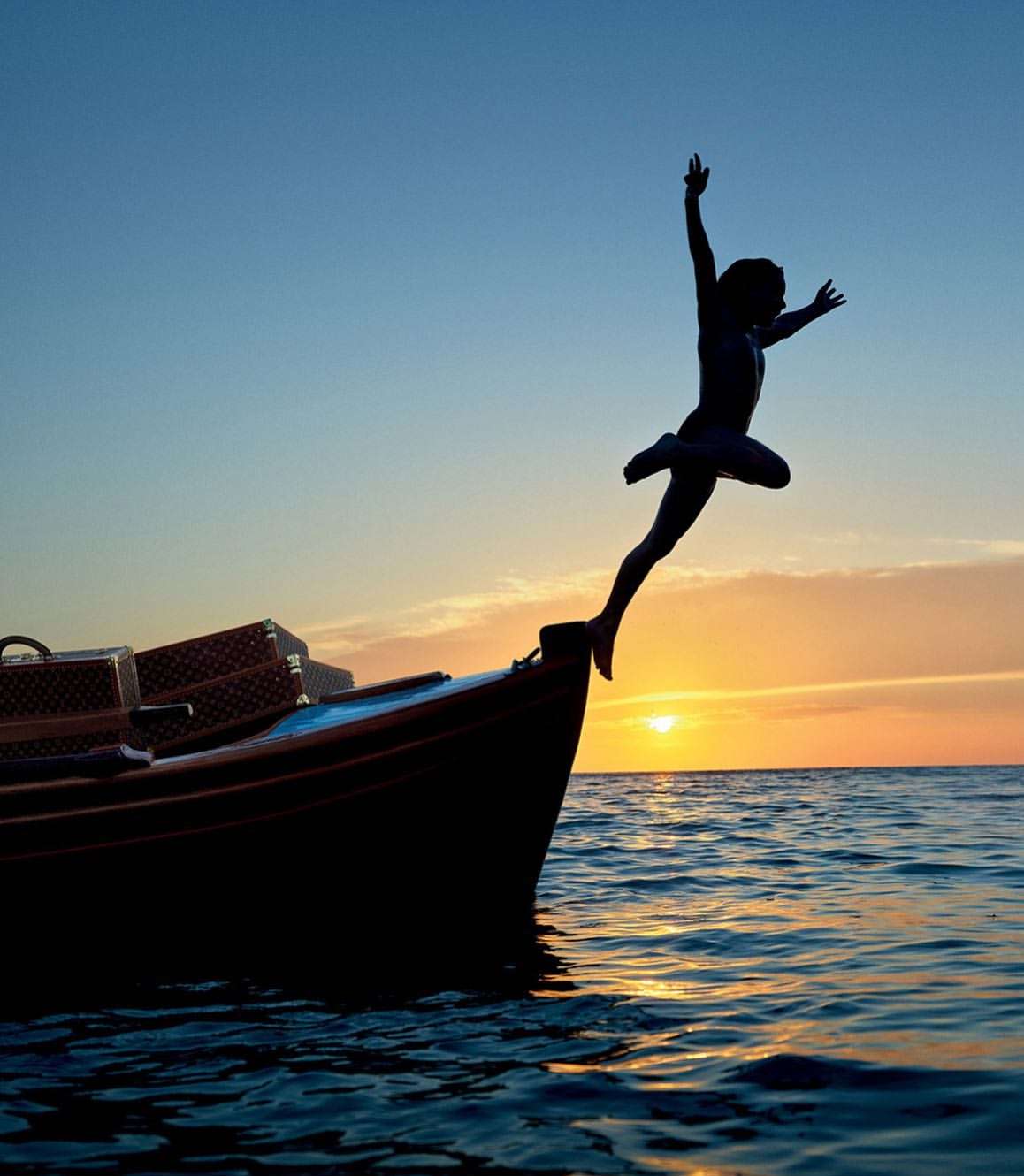 Silhouette of a person jumping from a boat into the ocean at sunset, with visible luggage on the boat and a clear sky in the background.