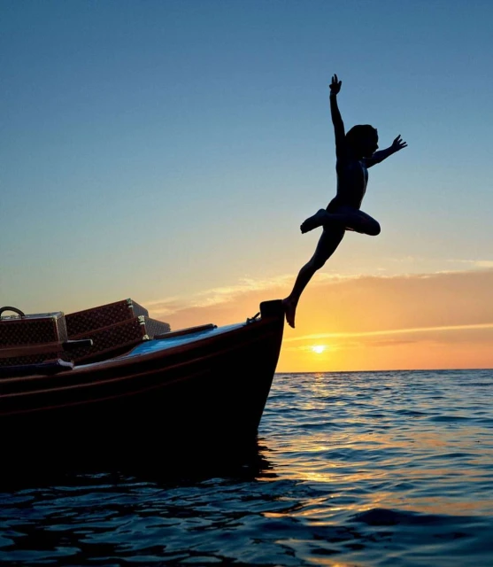 Silhouette of a person jumping from a boat into the ocean at sunset, with visible luggage on the boat and a clear sky in the background.