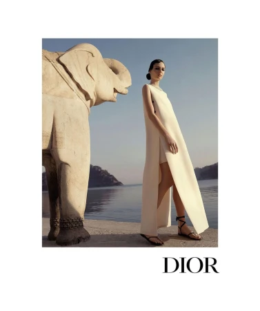 Fashion shot featuring a slender female model in an elegant white dress posing next to a stone elephant sculpture with a sea background.