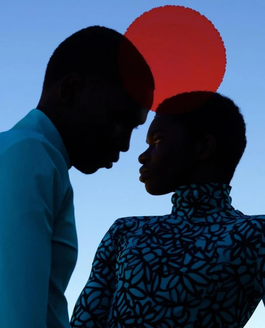 Two young Black adults stand close together in an outdoor portrait against a light blue sky with a red circular shape above them.