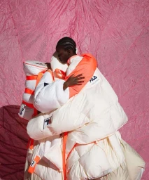 Two black women embracing while wearing bulky white and orange puffer jackets against a textured pink background