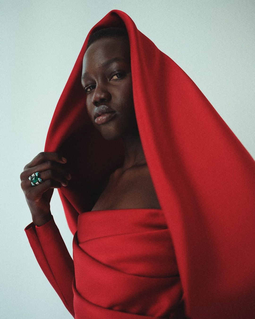 Woman in a red draped gown with a hood poses in front of a neutral backdrop, showcasing fashion and beauty in a portrait.