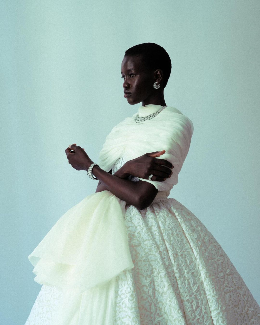 Elegant woman with dark skin poses in a white gown with a jeweled necklace, earrings, and bracelet against a light-blue backdrop.