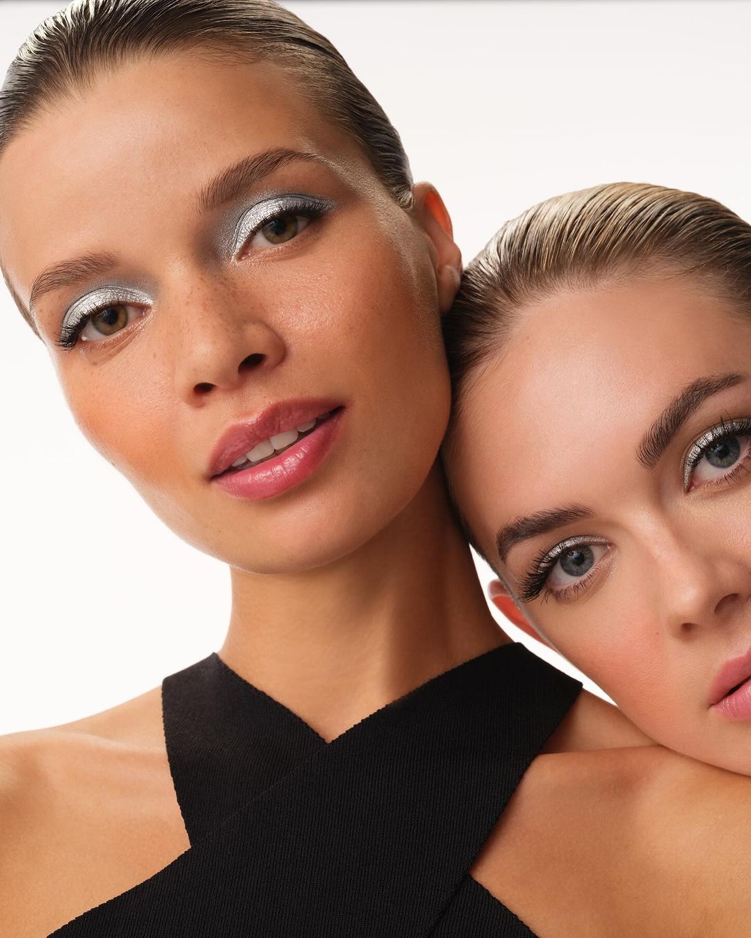 Close-up of two fair-skinned adult females with bold makeup, one resting head on the other's shoulder in a bright studio setting.