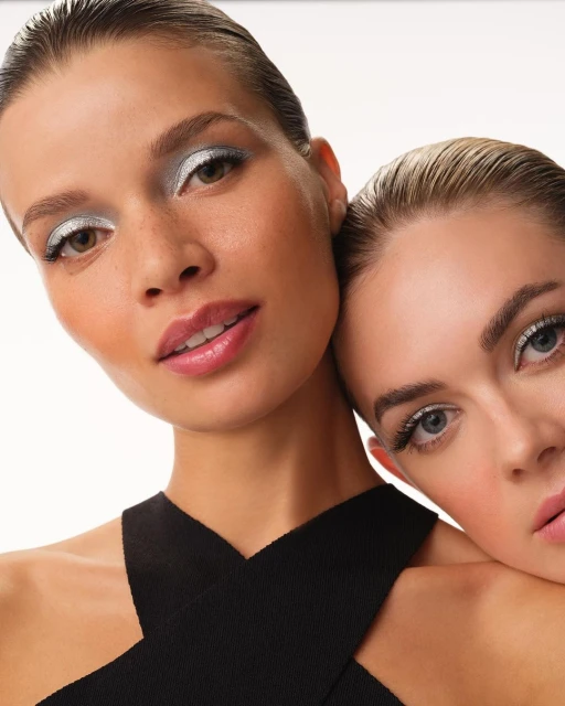 Close-up of two fair-skinned adult females with bold makeup, one resting head on the other's shoulder in a bright studio setting.