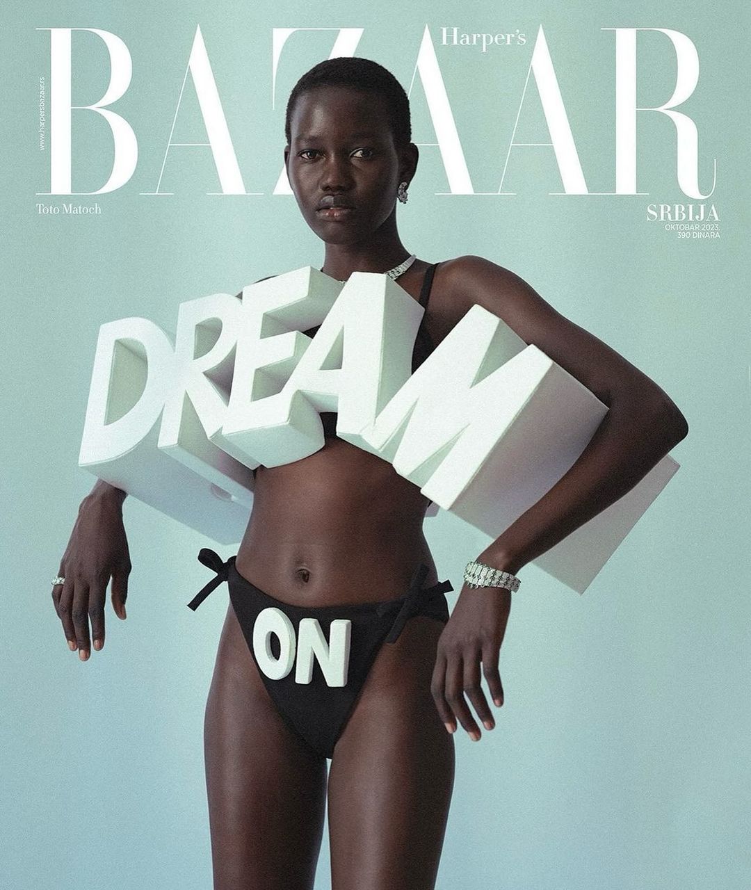 Editorial fashion shot of dark-skinned woman in black bikini bottom with 'ON' letters, holding DREAM letters, against a light-blue background.