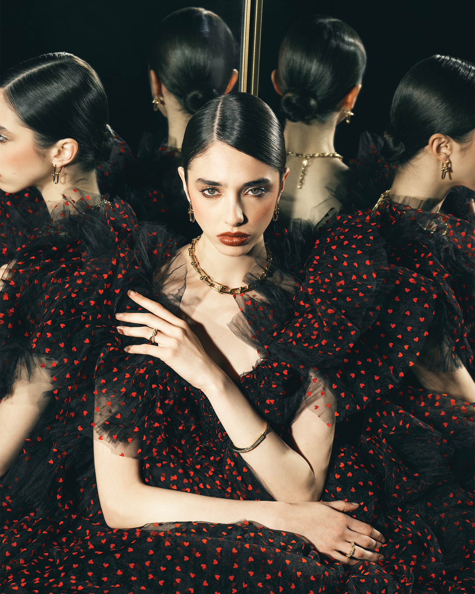 Portrait of a young woman with dark hair and red lipstick in a black dress with red heart patterns, accessorized with gold jewelry.