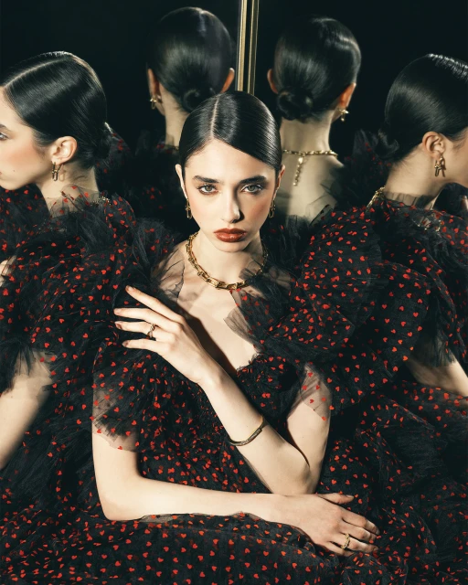 Portrait of a young woman with dark hair and red lipstick in a black dress with red heart patterns, accessorized with gold jewelry.