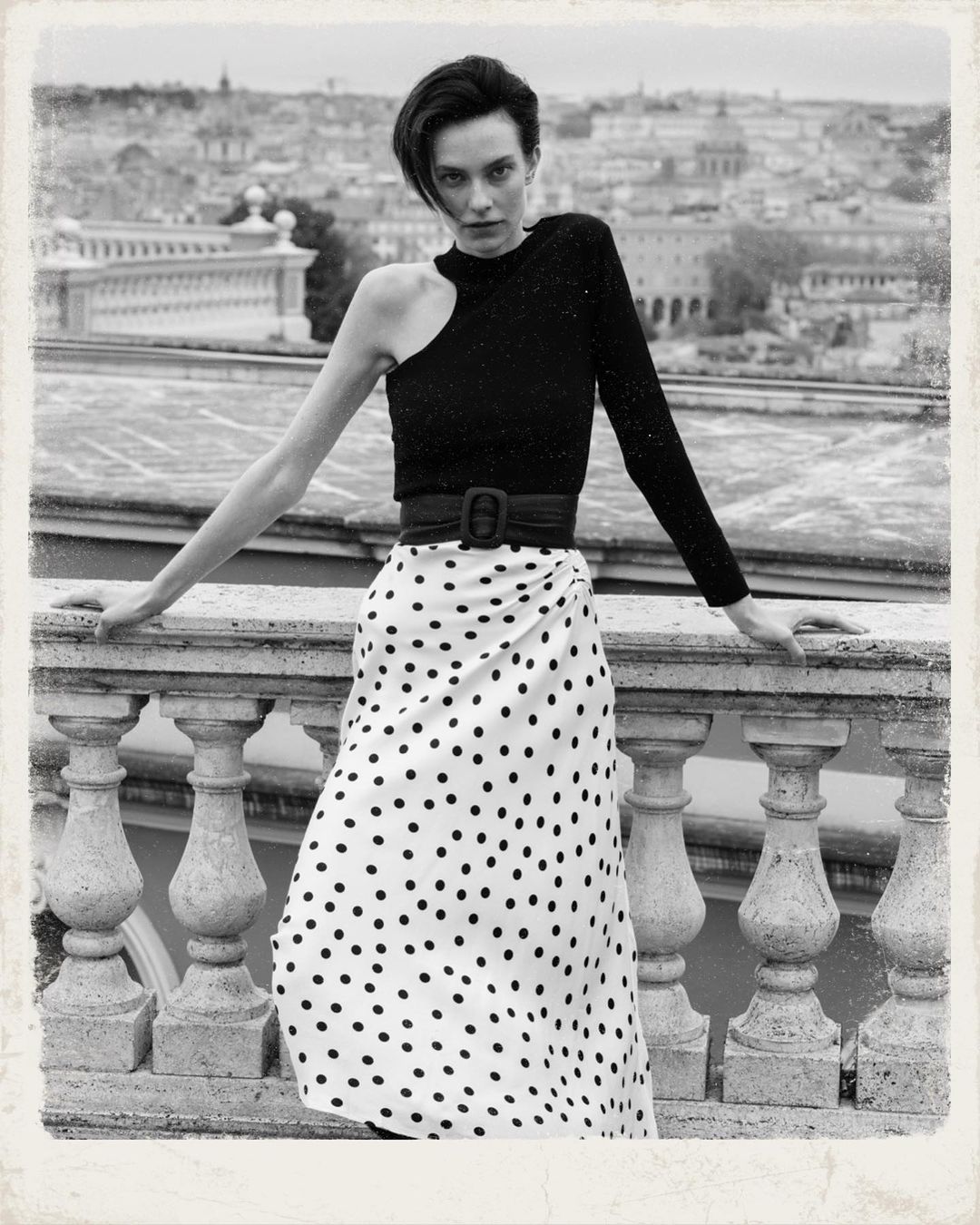 A fashion shot in black and white features a woman in a black top and polka dot skirt posing on a stone balcony with a city view.