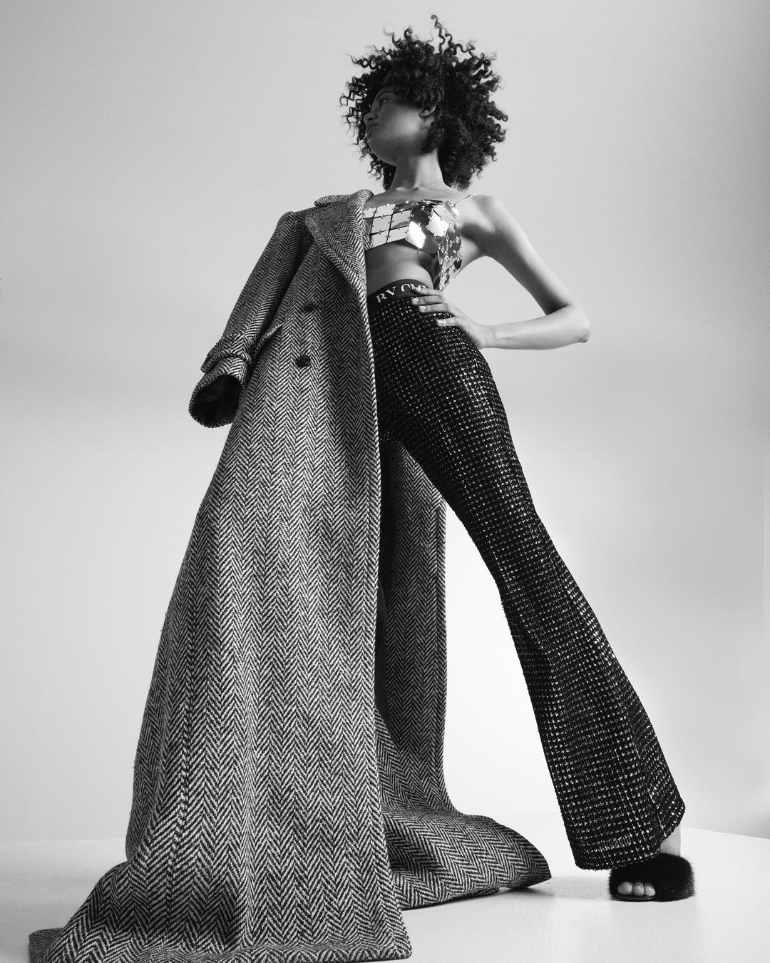 Black and white portrait of a woman wearing shiny pants and top with a coat hanging off her arm in a studio setting.