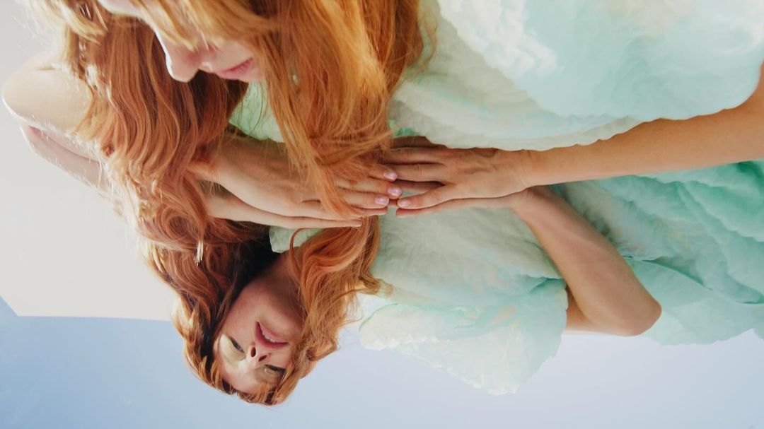 Close-up shot of a red-haired woman in a light-blue ruffled dress, reflected in a mirror or reflective surface, showcasing a serene composition.
