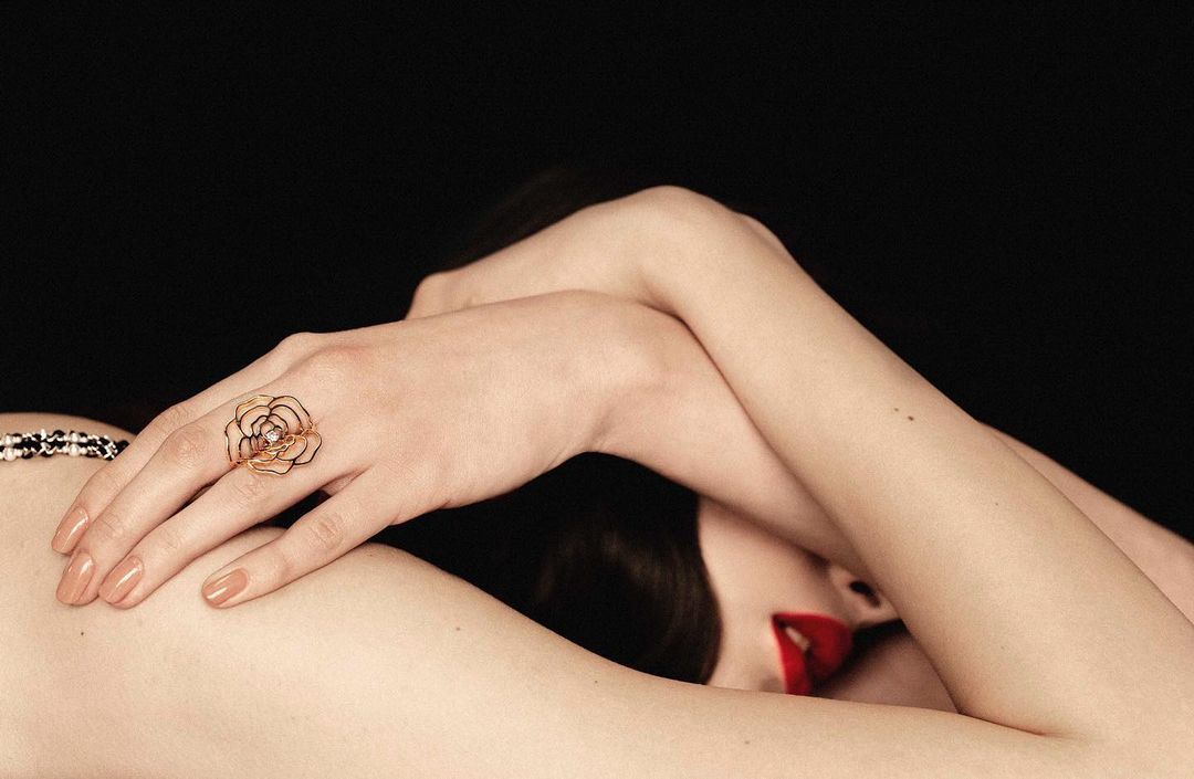 Close-up of a woman lying down wearing a gold-tone, floral ring, glossy red lipstick, a beaded top and posing in a black studio.