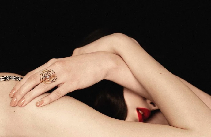 Close-up of a person resting their head on crossed arms, displaying an intricate gold ring on the hand and vibrant red lipstick.