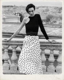 Model in black one-shoulder top and polka dot skirt leaning on rooftop balustrade with cityscape background