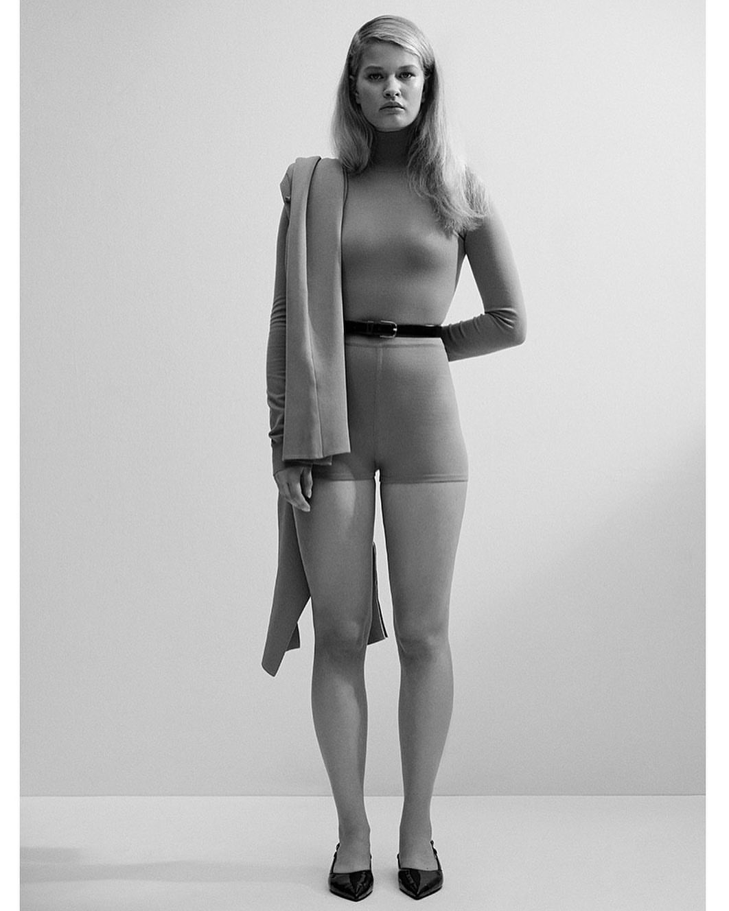 Full-length studio shot of a blonde female model wearing a matching top and shorts, black belt, and black heels with a jacket draped over her shoulder.