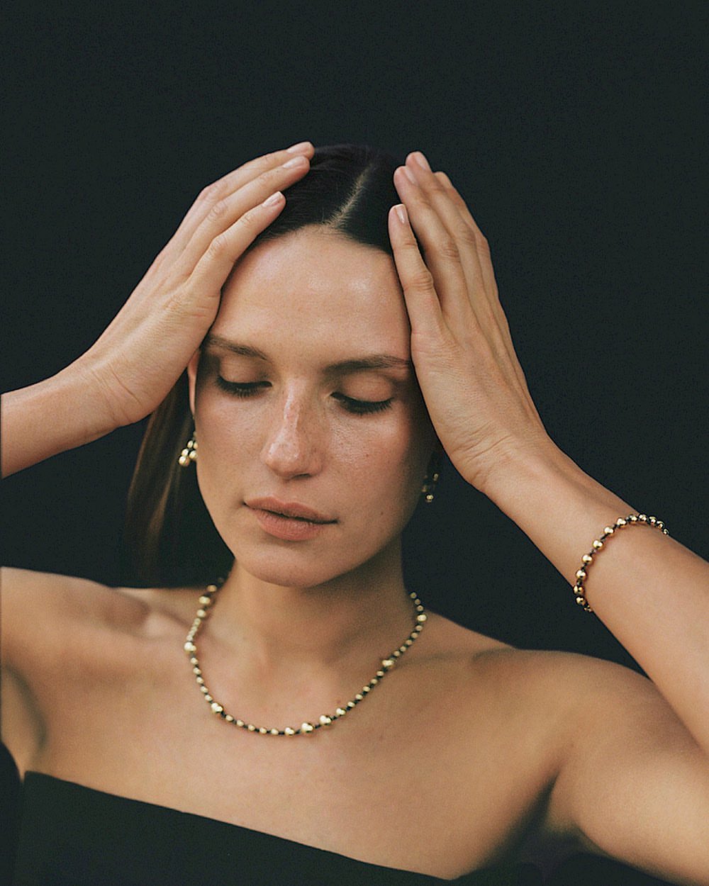 Portrait of a fair-skinned adult female in jewelry posing indoors against a dark background with her hands near her head.