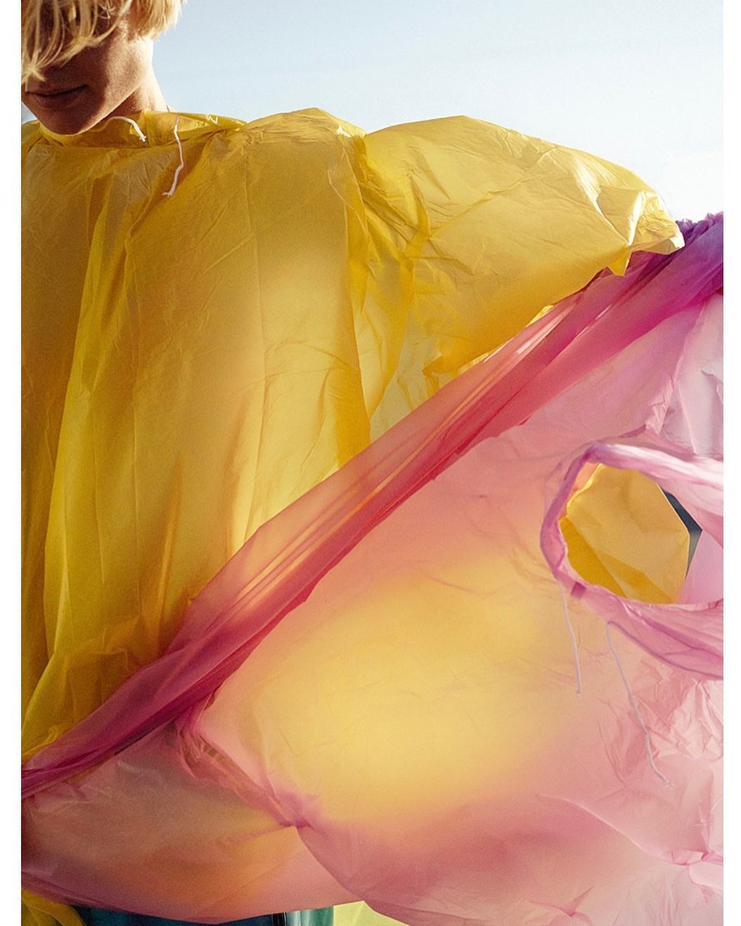 Person in yellow plastic garment with red cellophane, illuminated by sunlight, against a clear blue sky, in a vertical composition.