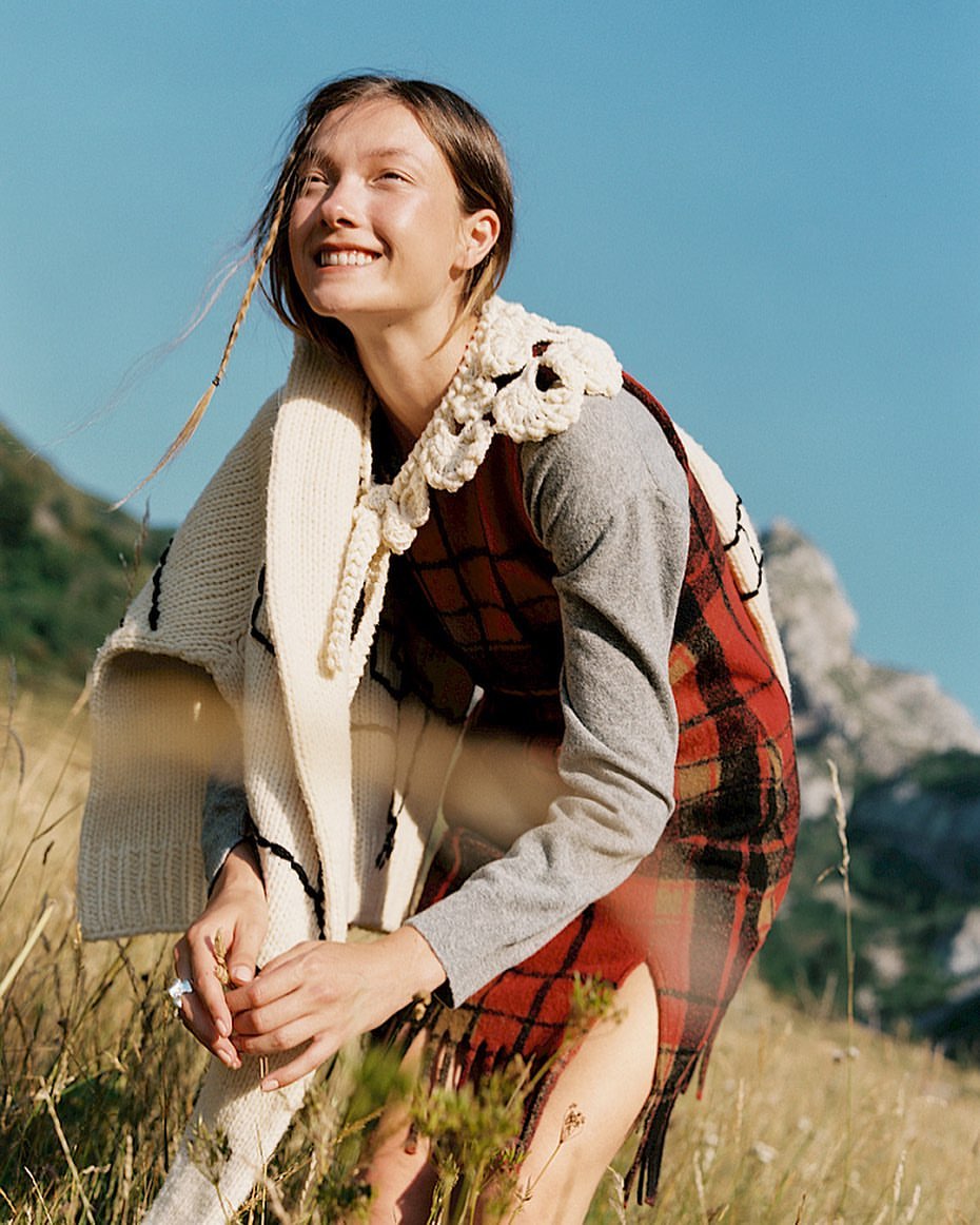 Smiling young woman wearing a red plaid dress and cream-colored crochet shawl in a sunlit, grassy outdoor environment with mountains.