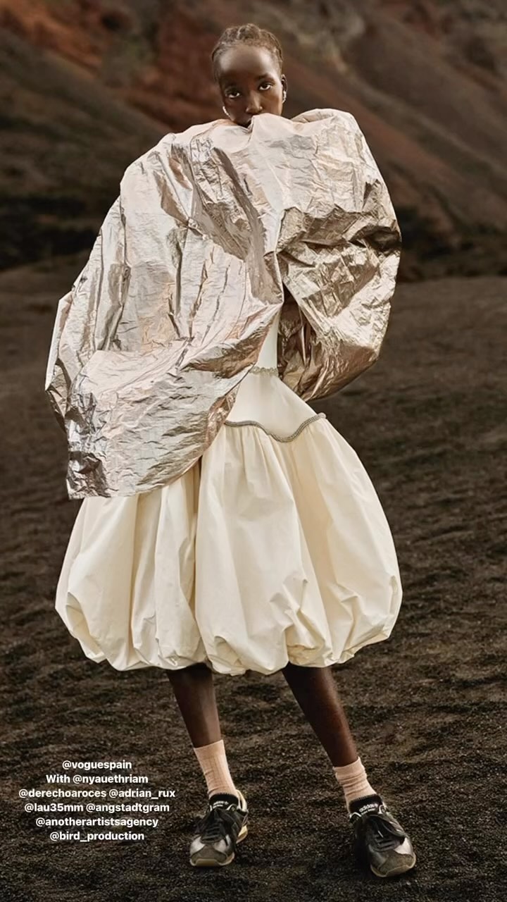 Fashion editorial shot of Black woman in puffy white dress and silver metallic jacket posing outdoors on black ground with brown mountains.