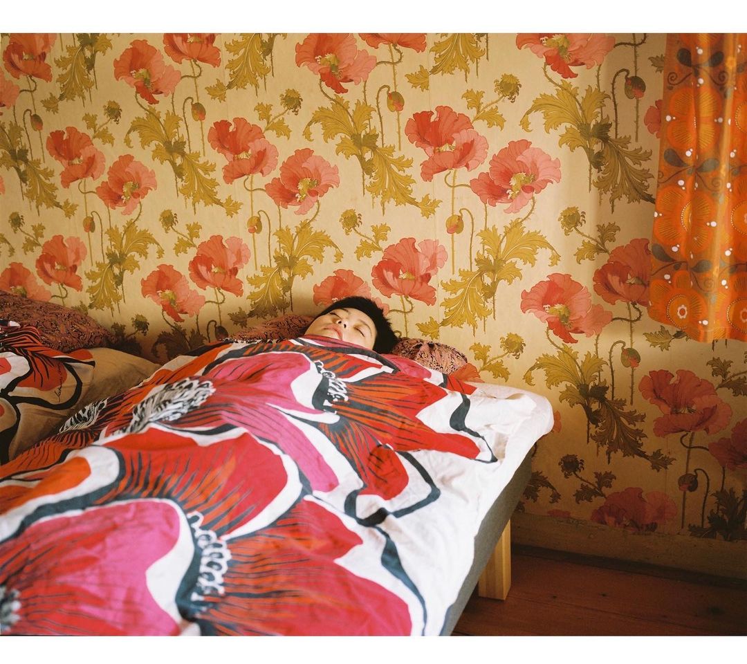 A young person rests in a bed covered by a bright floral blanket, set against a wall decorated with a matching poppy wallpaper.