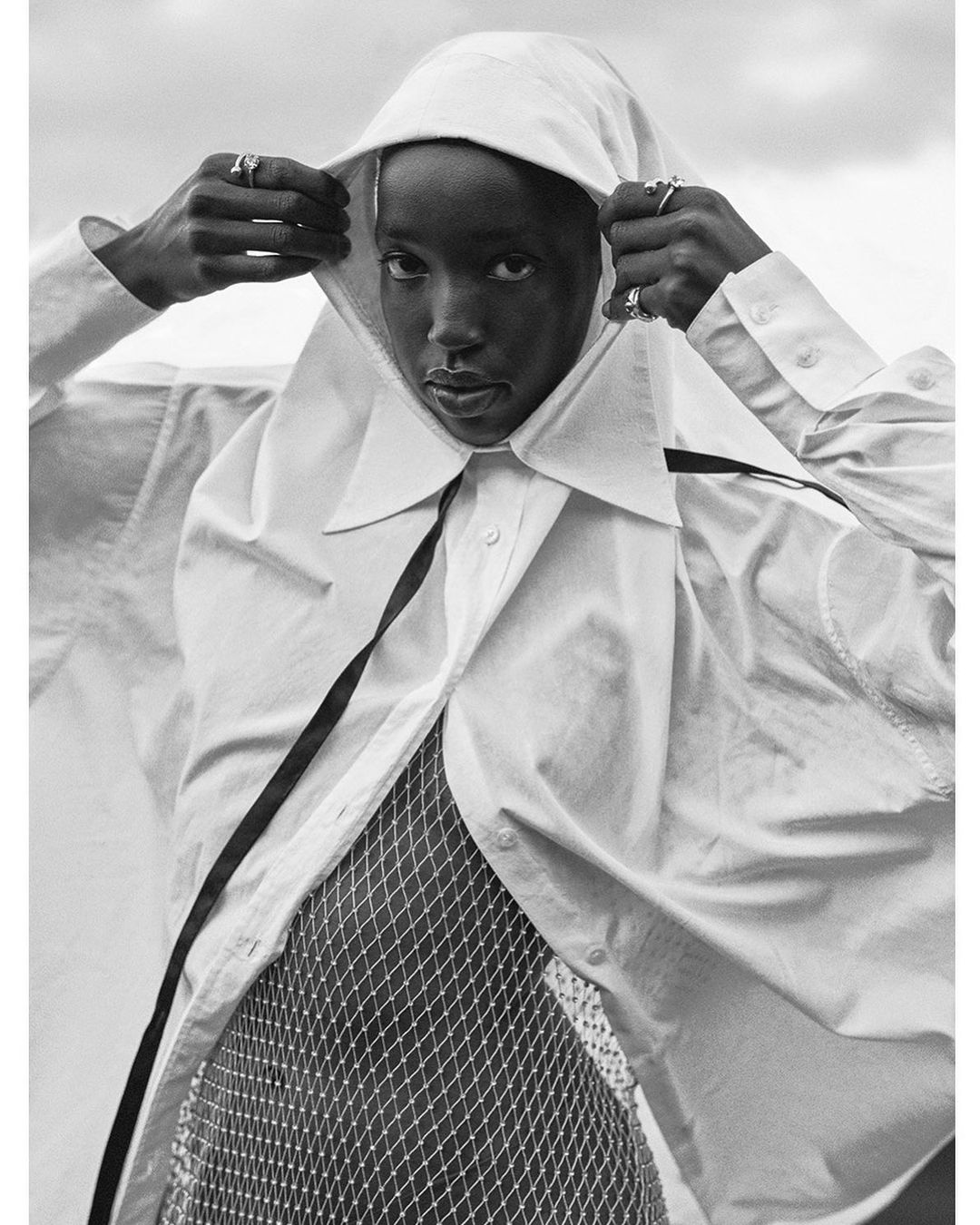 Striking black and white portrait of a woman with hands framing a hood over her head, wearing a white shirt and mesh top.