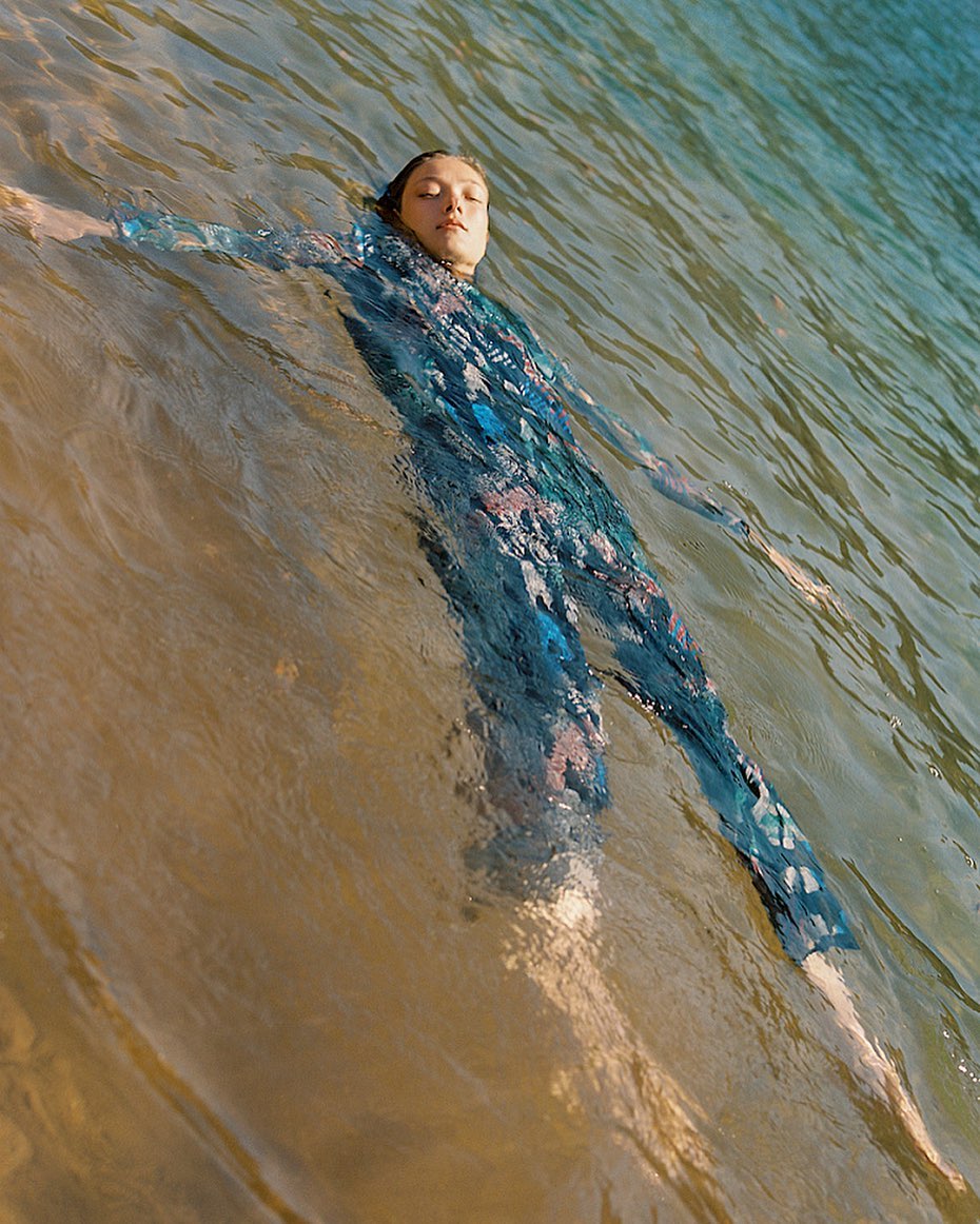 Woman in a blue patterned full-body swimsuit, lies floating in clear water with serene expression, wet hair, in an eye-level outdoor shot.