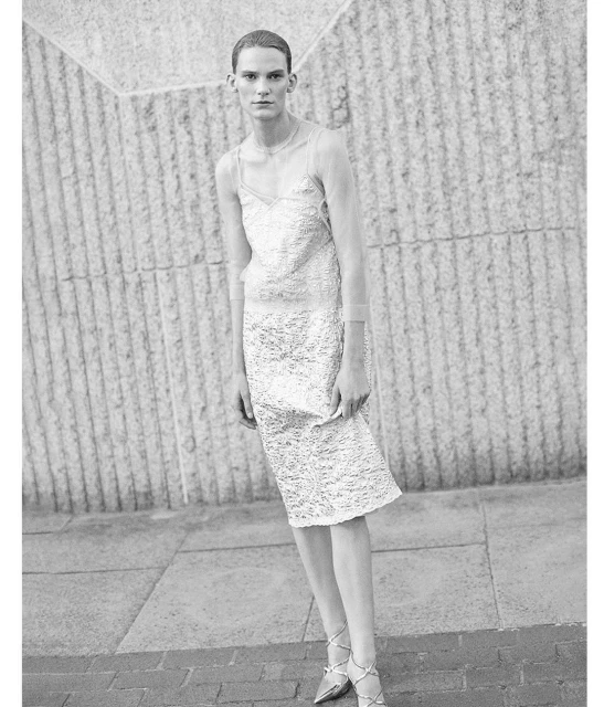 A woman in a light-colored dress and heels poses in front of a textured wall, in a black and white fashion shot with soft lighting.