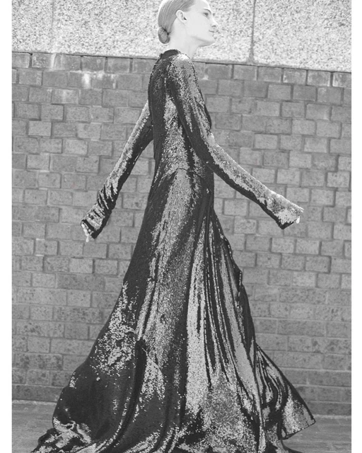 Woman in long-sleeved sequin gown walks against a textured wall in this black-and-white shot highlighting fashion and style.