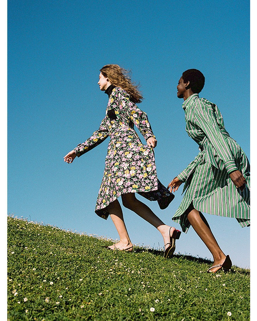 Two women in dresses walking up a grassy hill against a blue sky. One has a floral dress; the other has a green-striped dress.