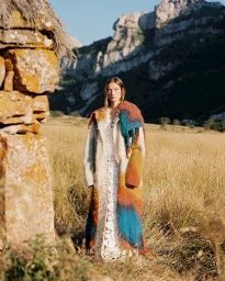 Model wearing a long textured colorful coat over a white lace dress standing in a grassy meadow beneath mountains.