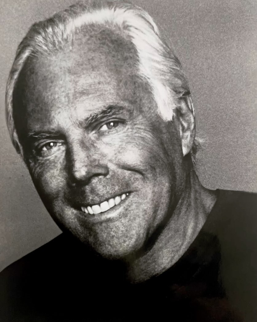 Portrait of Giorgio Armani with white hair and dark t-shirt smiling against a textured grey background.