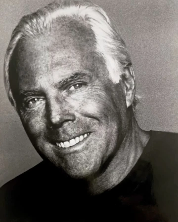 Portrait of Giorgio Armani with white hair and dark t-shirt smiling against a textured grey background.