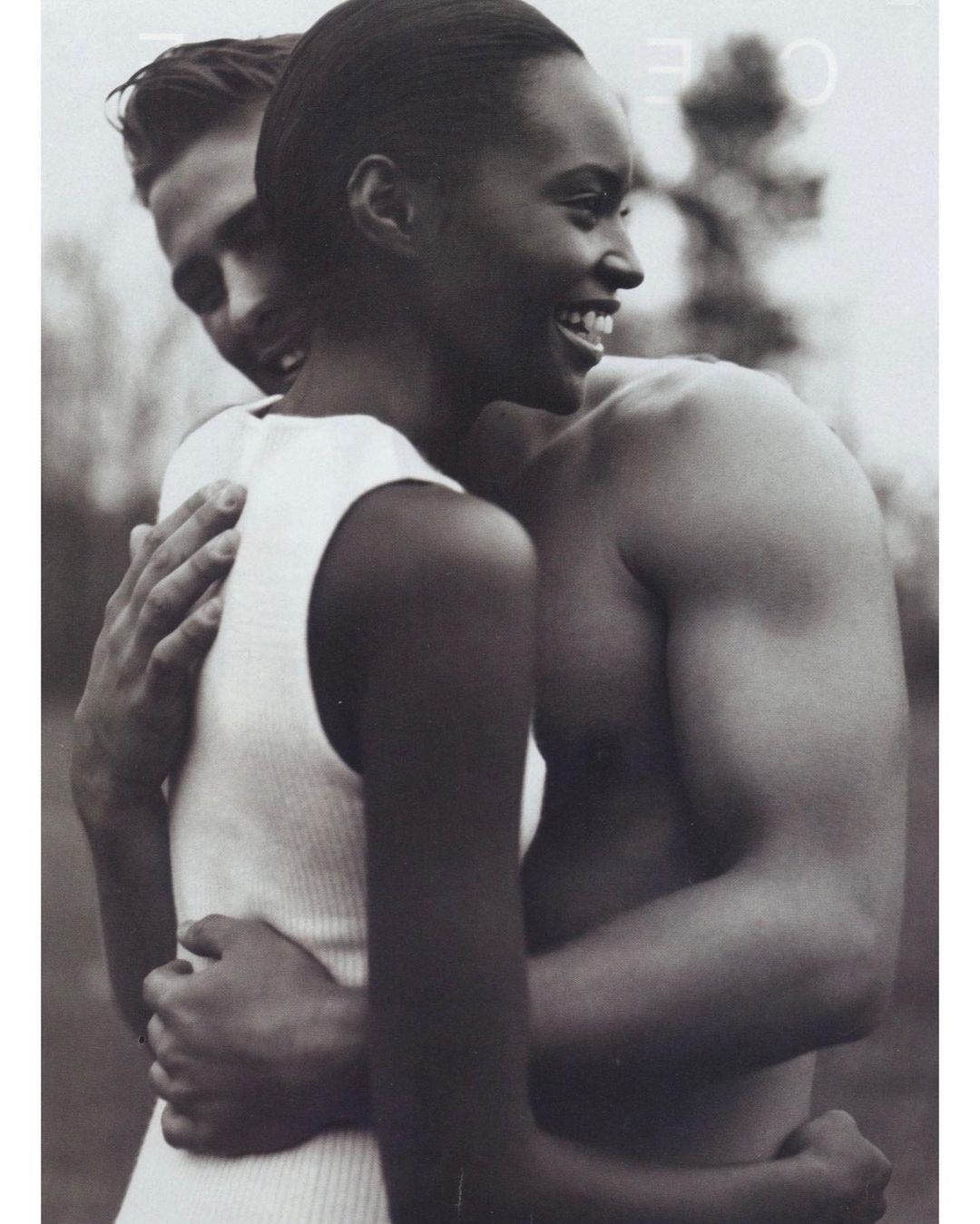 Black and white studio portrait of a smiling Black woman being embraced by a shirtless Caucasian man against a blurred background.