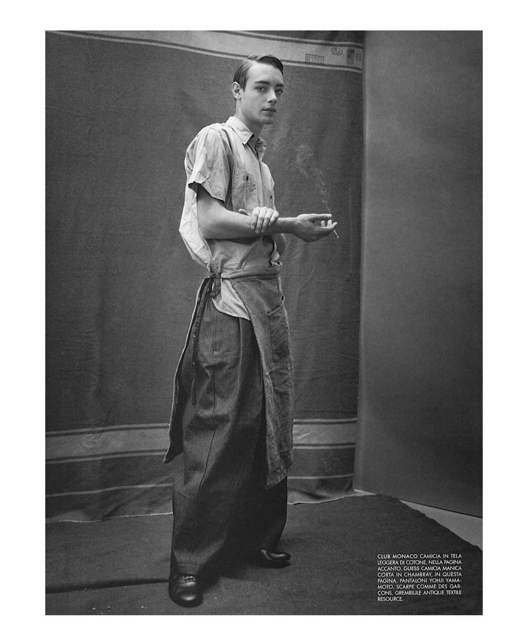 Monochrome portrait of a young man wearing casual clothes and a denim apron, holding a cigarette, against a neutral backdrop.