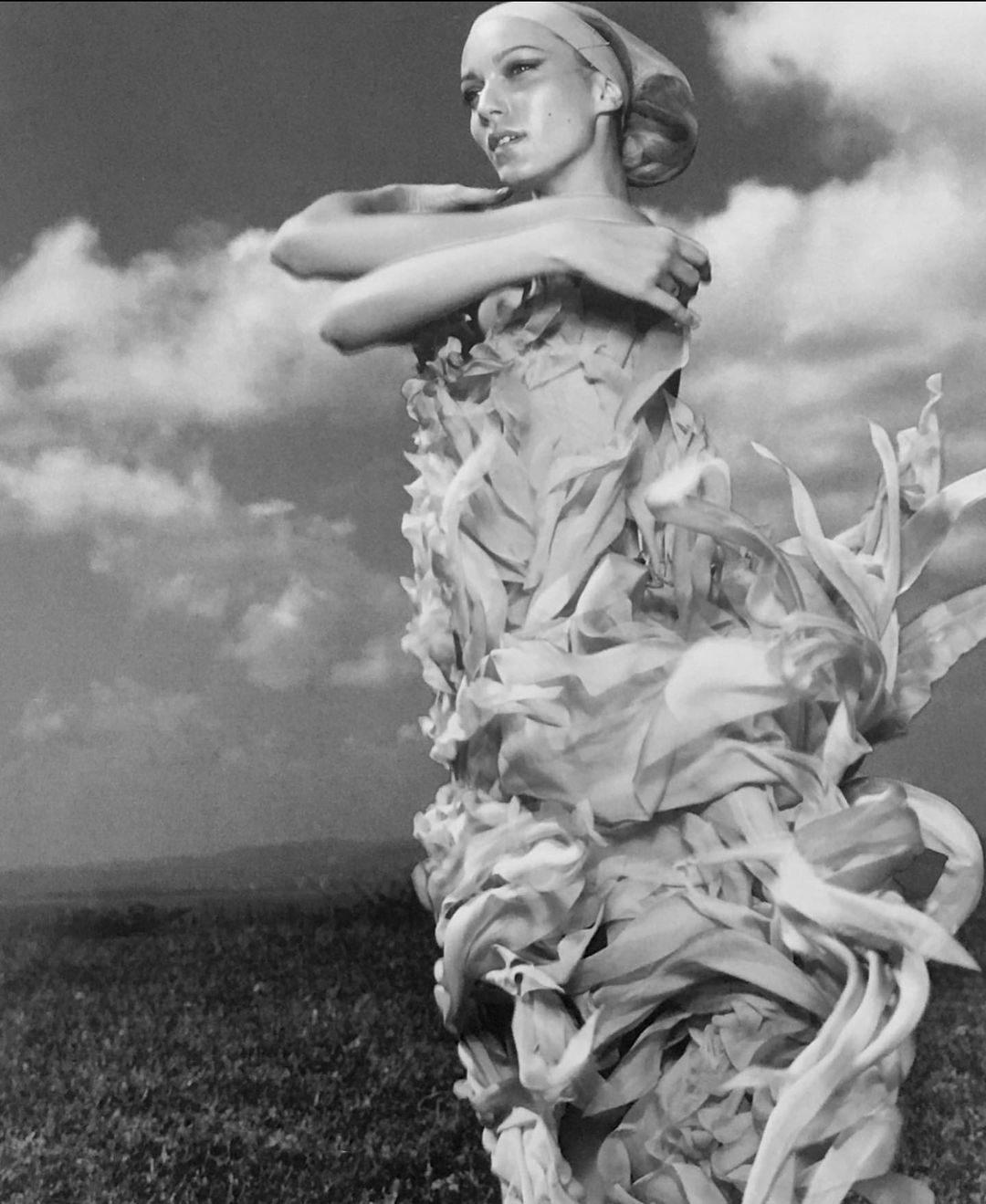 Woman in an artistic dress posing in a field under a cloudy sky. The black and white photograph showcases a unique blend of fashion and nature.