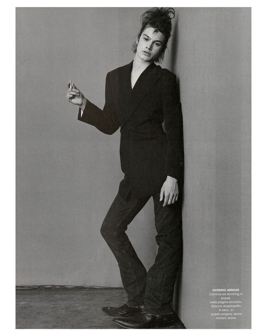 Fashion editorial shot of a young adult leaning against a wall, wearing a dark suit with messy hair, and holding a cigarette indoors.