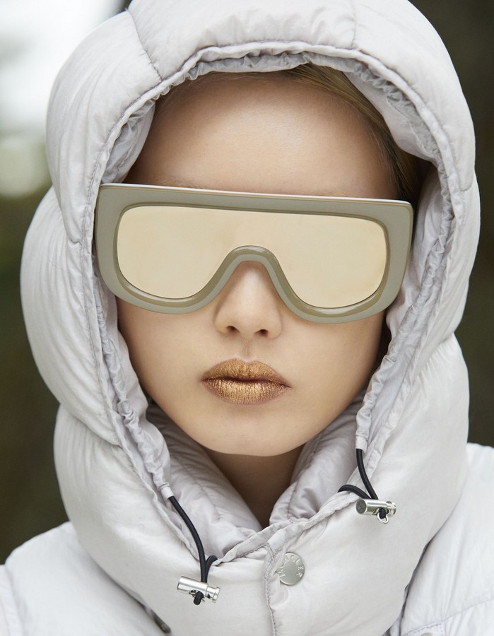Close-up of a model in an off-white puffer jacket with the hood up, large square sunglasses with reflective lenses, and gold lipstick.