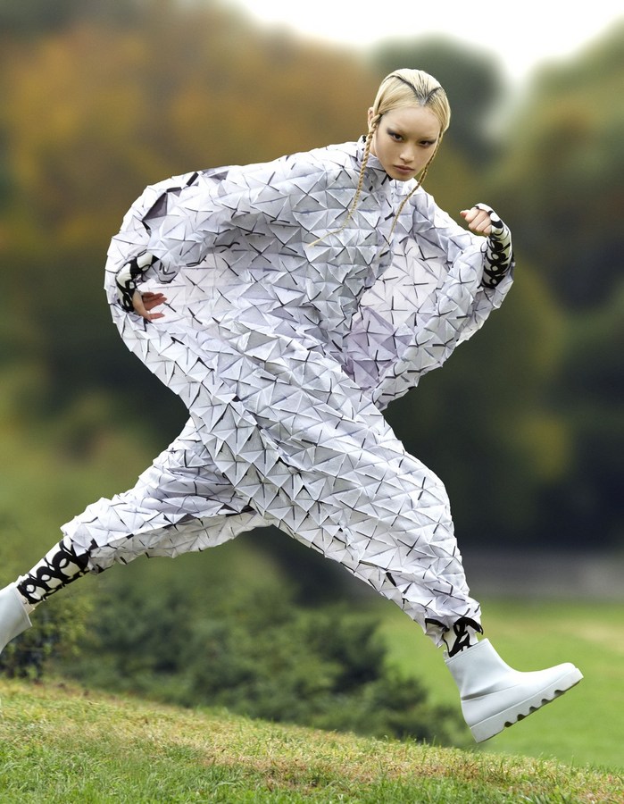 Woman in futuristic geometric outfit leaps across grassy hill. Outdoor fashion shot with trees in the blurred background. Unique style.