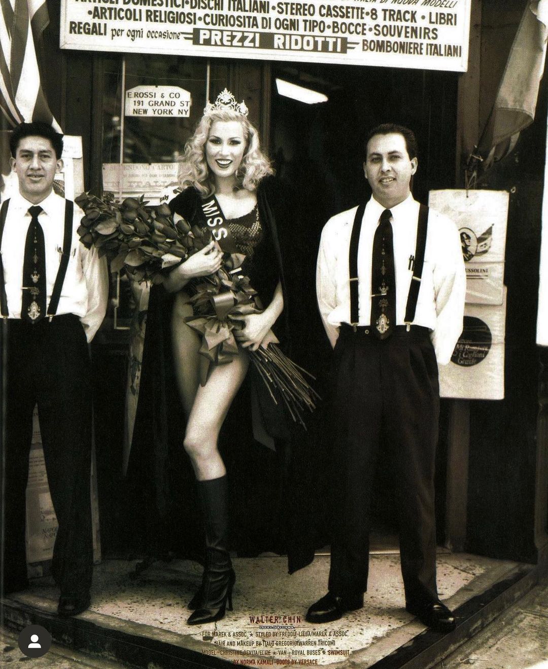 Pageant winner stands with bouquet, crown, and cape between two men in front of Italian goods store, medium shot, black and white photo.