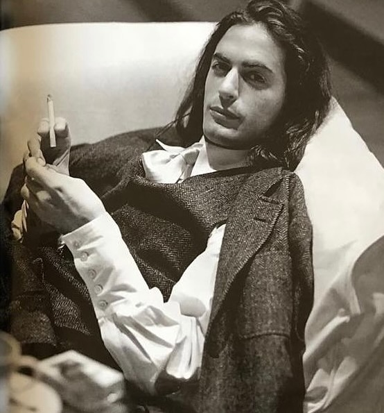 Elegant man with long hair lounges with cigarette in a herringbone suit, posing in a brightly lit, vintage-style indoor setting.