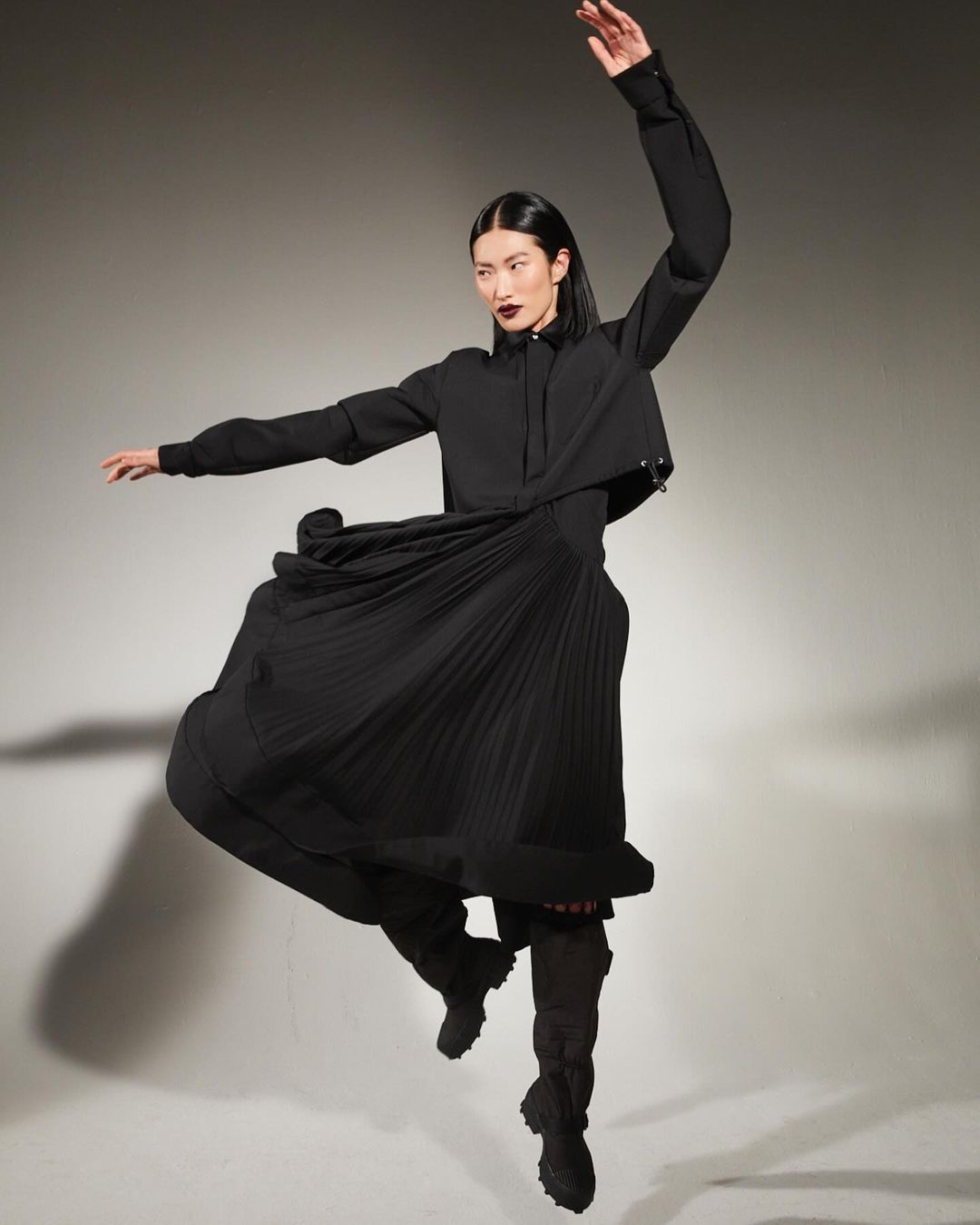 A woman in a black long-sleeve shirt, skirt, and boots leaps in a studio with arms spread. Fashion shot in a neutral studio backdrop.