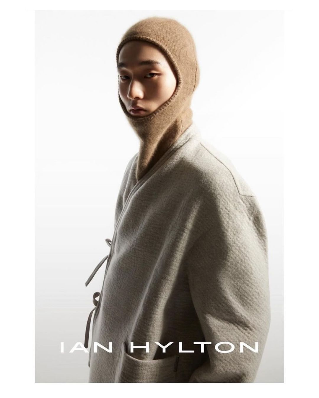 Portrait of a young adult wearing a brown knit balaclava and textured neutral-tone jacket against a bright white studio backdrop.
