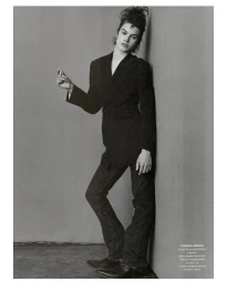 Model in black jacket and dark trousers leaning against a wall holding lipstick, photographed by Walter Chin in New York.