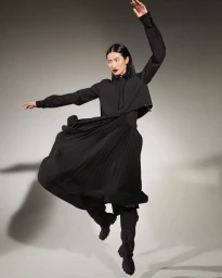 Model in long black pleated dress and dark boots leaping against a grey studio seamless background by Walter Chin.